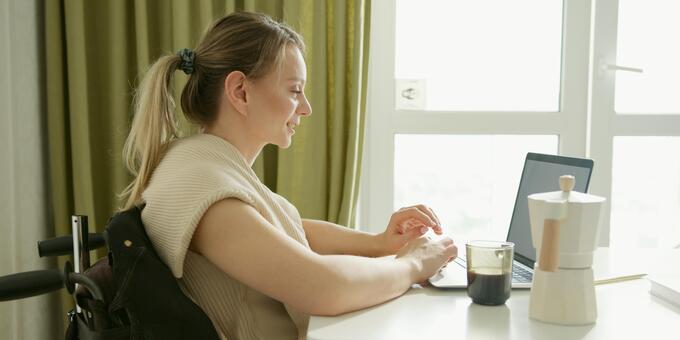 Eine junge Frau sitzt am Tisch in einem Rollstuhl. Sie nutzt einen Laptop.