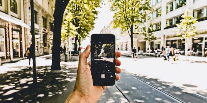 Eine Person ist in einer Stadt unterwegs und hält ein Smartphone. Die Person macht gerade ein Foto der Straße. Es sind Straßenbahnschienen, Gebäude und Bäume zu sehen.