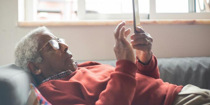 Ein Senior liegt auf dem Sofa und liest etwas am Smartphone.