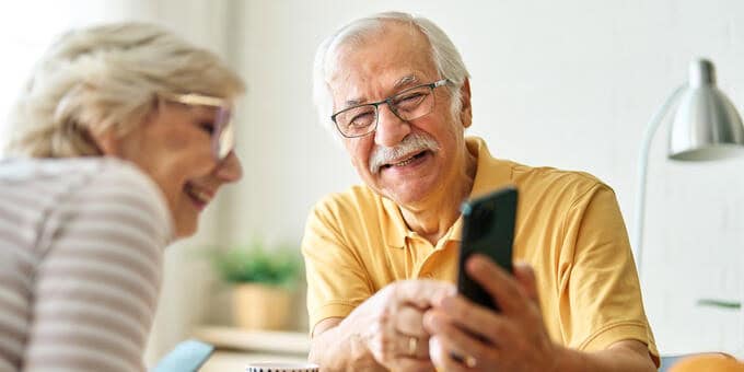 Zwei ältere Personen sitzen am Tisch. Der ältere Herr zeigt der älteren Dame etwas auf dem Handy und beide schauen fröhlich.